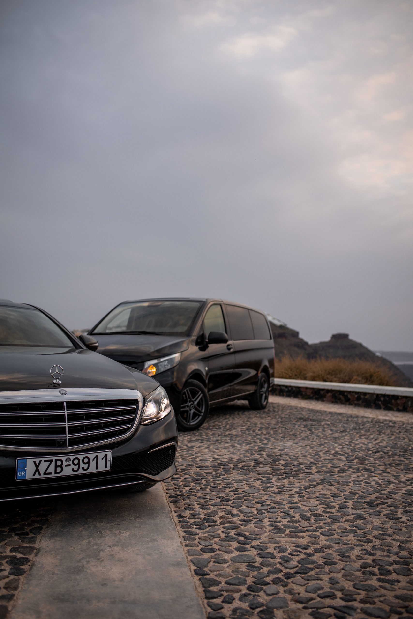 Black Mercedes-Benz E-Class and Vito van parked on a cobbled clifftop road in Santorini