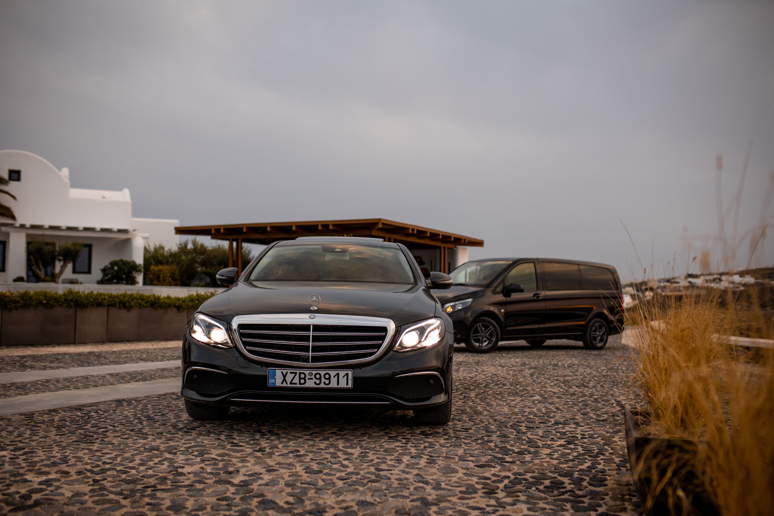 Mercedes-Benz E-Class with headlights on and a Vito van parked by a Santorini villa