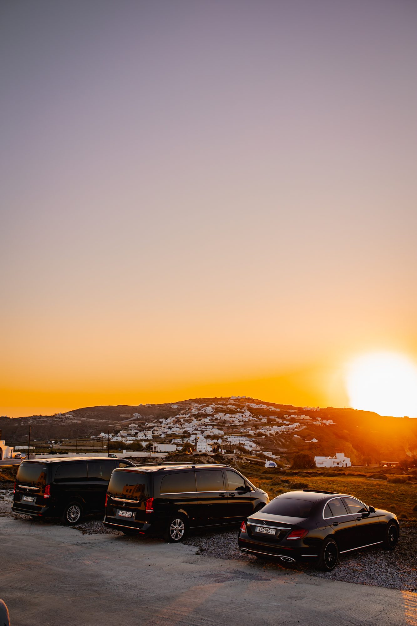 Three Mercedes-Benz transfer vehicles parked at a Santorini viewpoint during golden-hour sunset