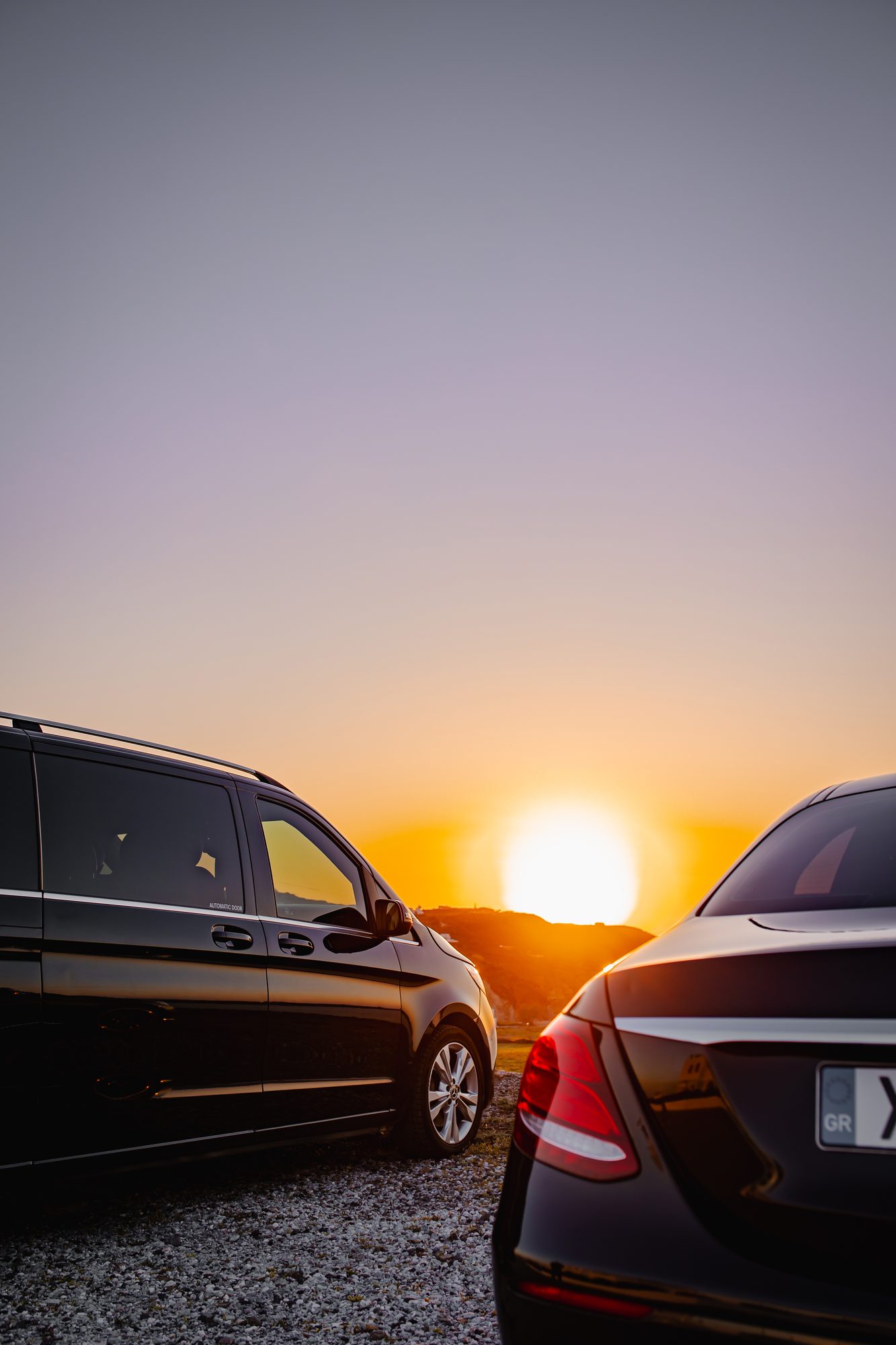 Mercedes-Benz V-Class van and E-Class sedan framing the sun setting over the Santorini hills