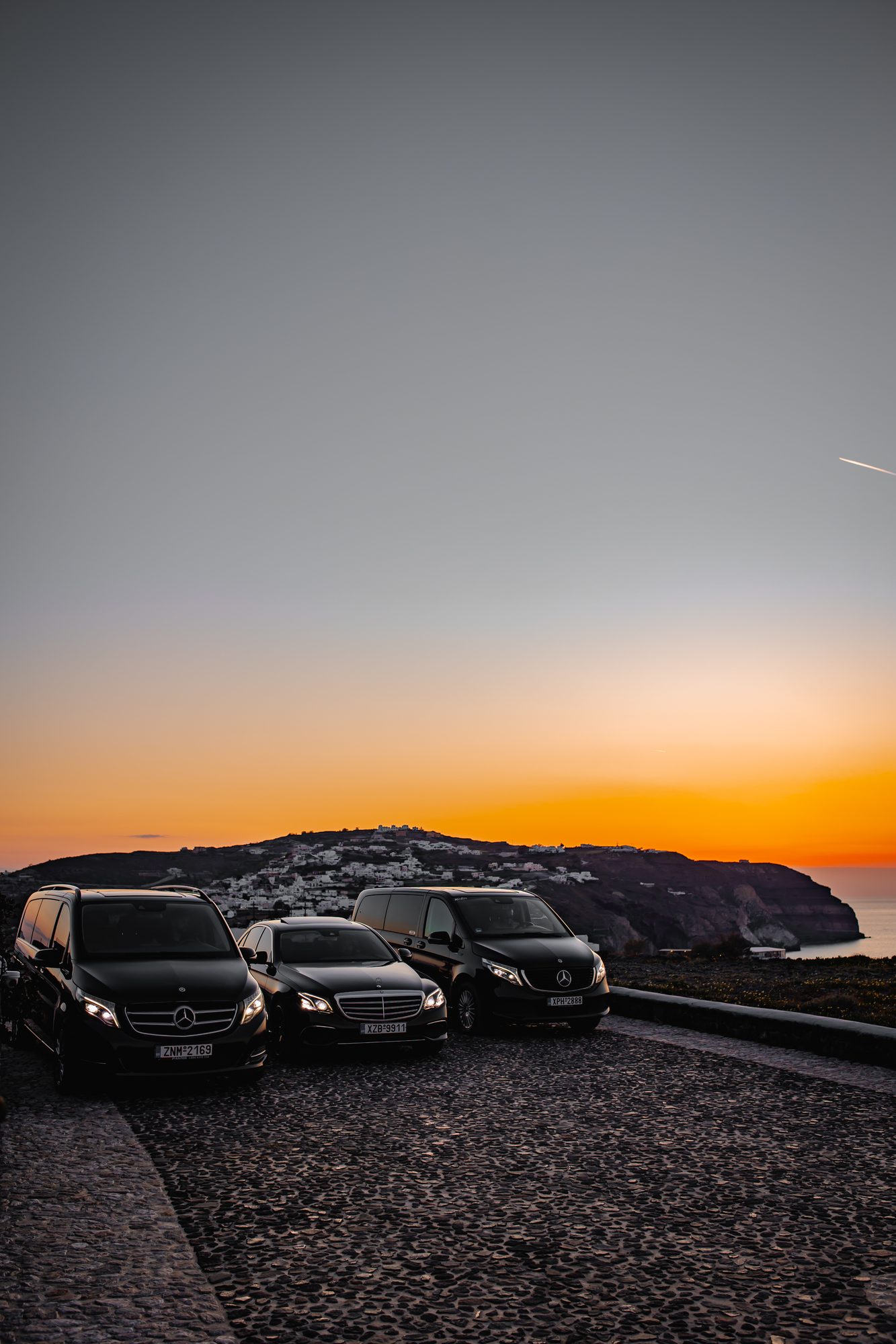Mercedes-Benz fleet parked above the caldera with a Santorini village glowing at sunset